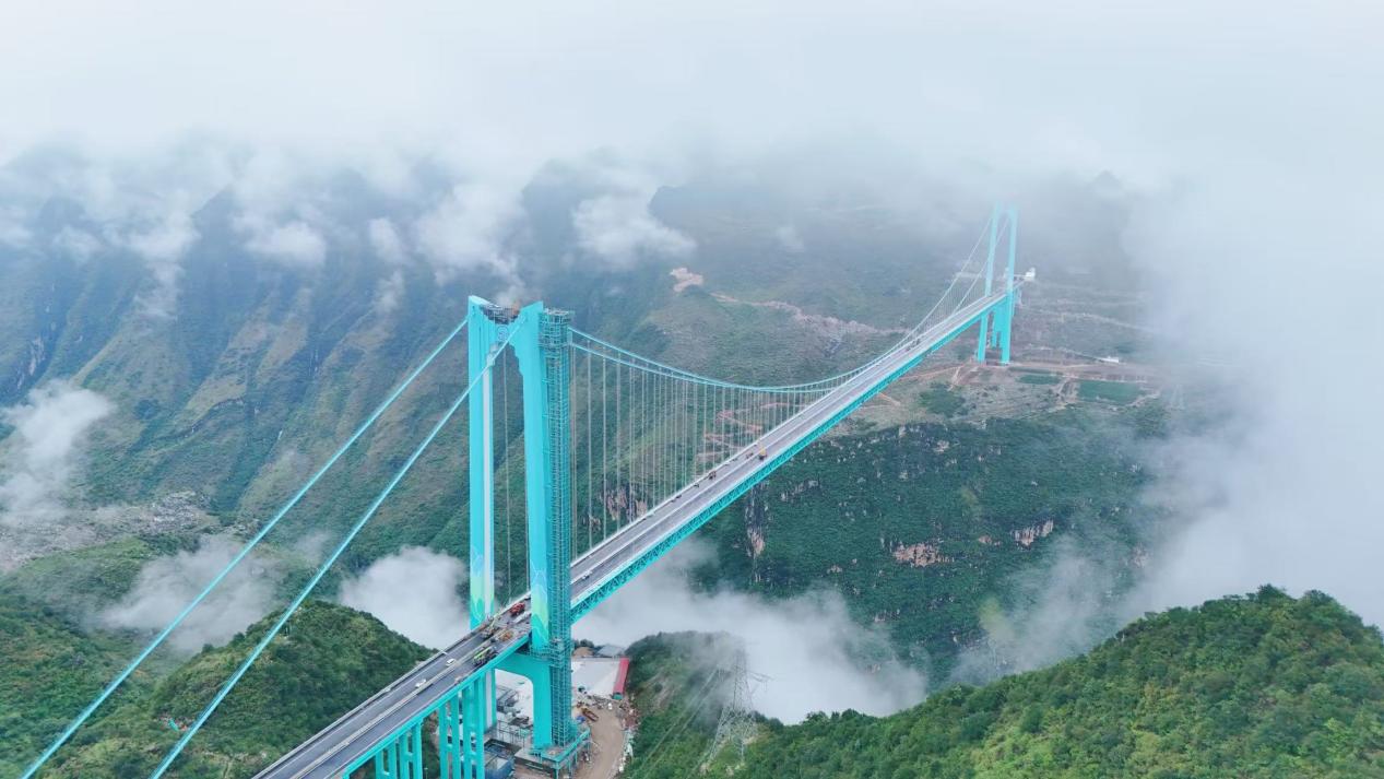 A massive, bright blue suspension bridge with the longest span in the world stretches across misty green mountains, showcasing advanced engineering systems like the智慧缆索吊装系统 and多普勒激光雷达测风系统.